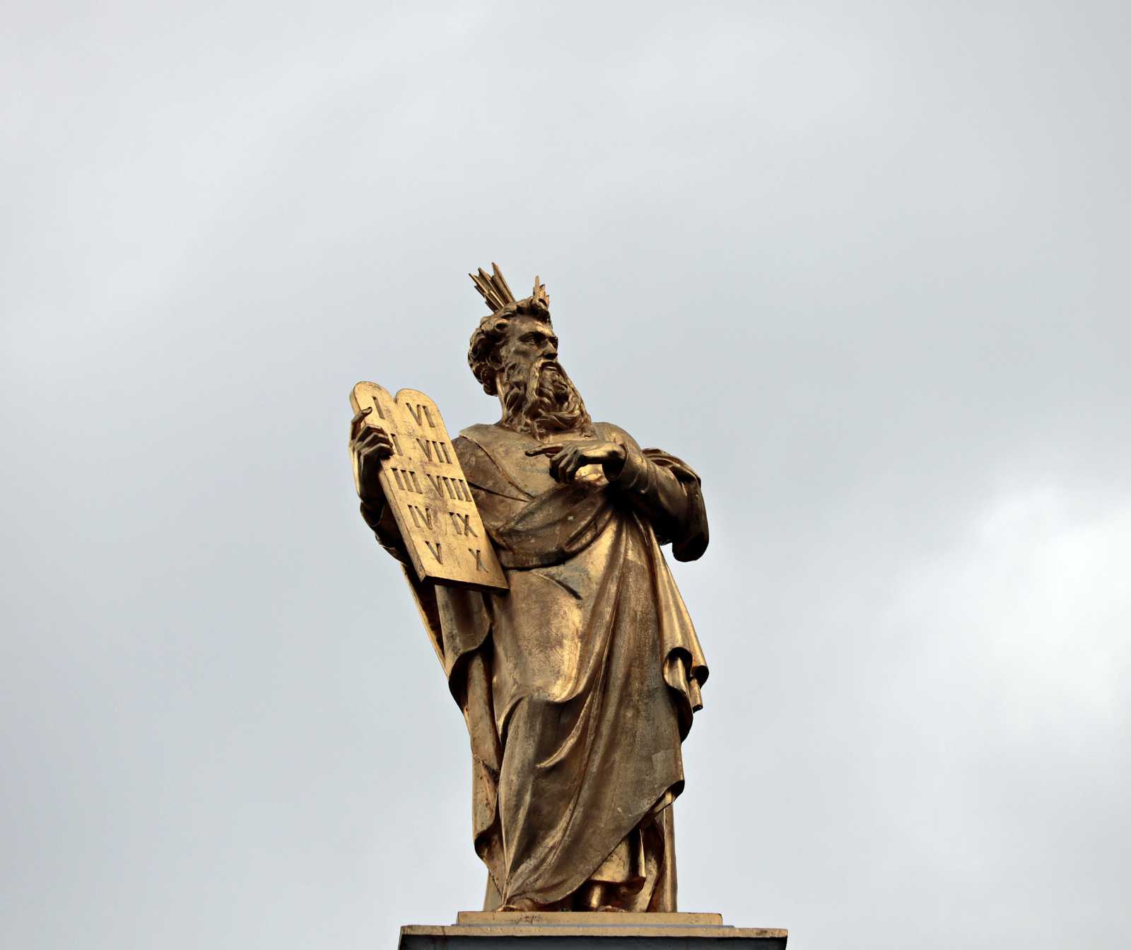 Goldene Statue eines Mannes mit Umhang und einem Kopfschmuck. Er hält eine Tafel mit den römischen ziffern ein bis zehn darauf in der Hand