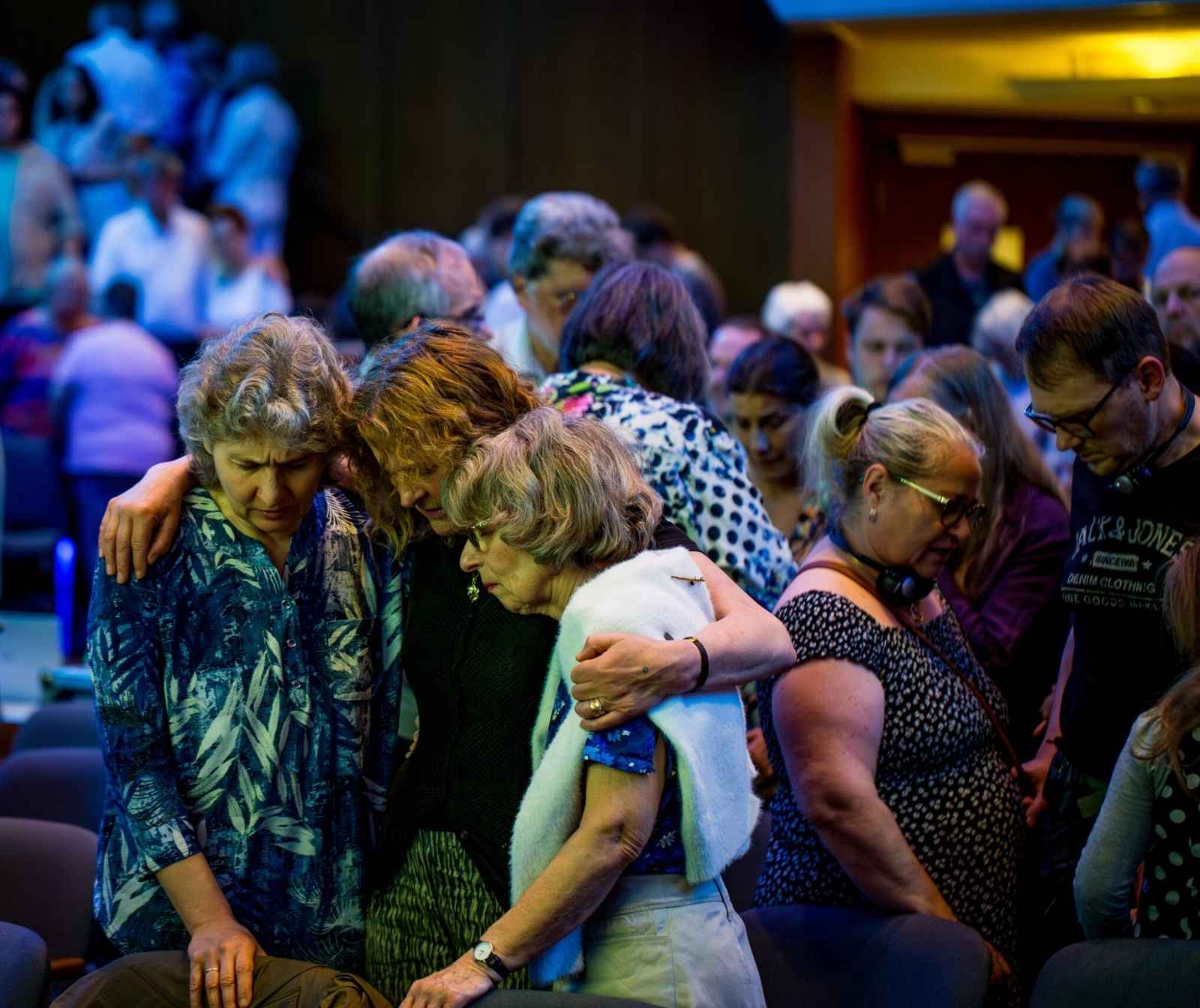in einem bunt ausgeleuchteten Saal stehen Menschen in Gruppen zusammen, Augen geschlossen und Kopf gesenkt. Manche haben ihre Arme über die Schultern anderer gelegt