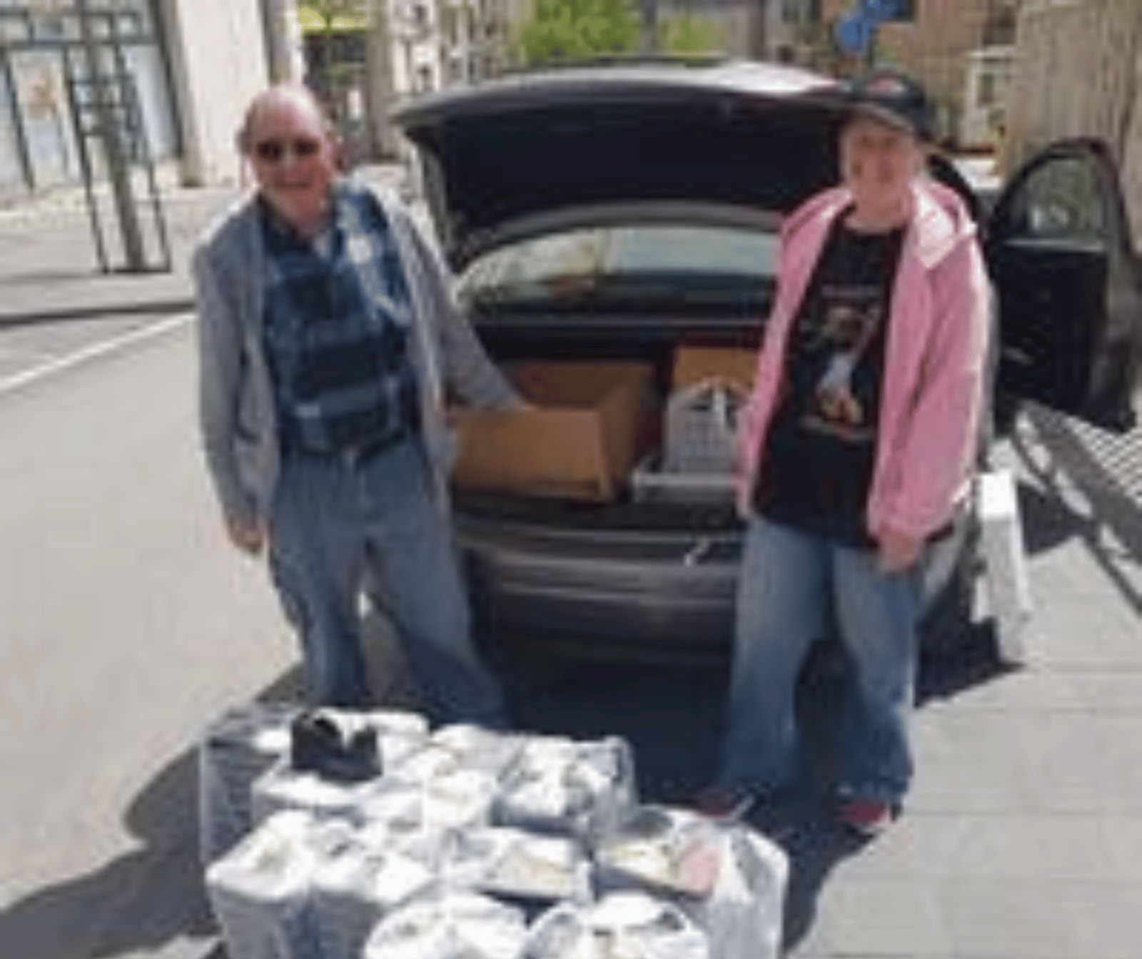 zwei Menschen vor einem geöffnetem Kofferraum befüllt mit Kartons. Vor ihnen stehen weitere Päckchen auf dm Boden