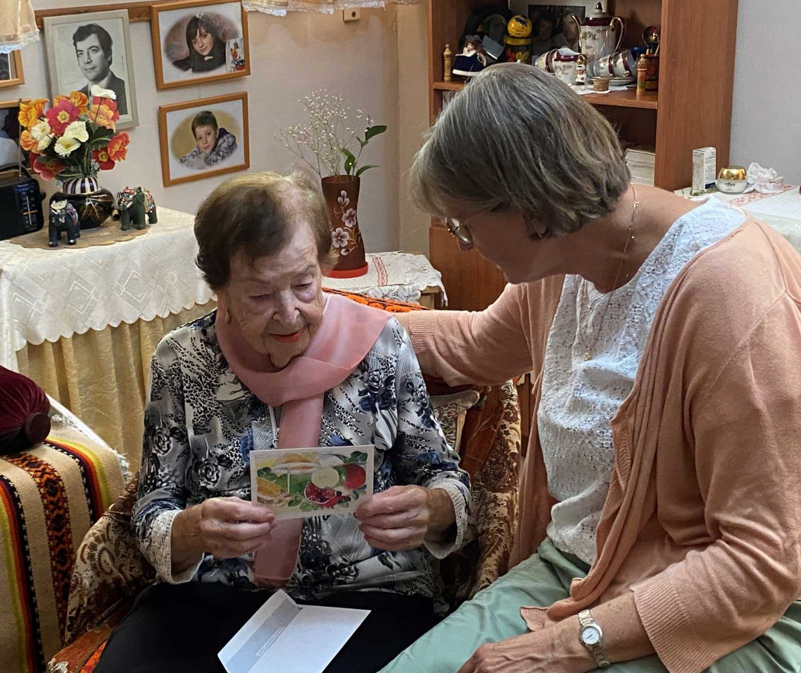 eine Frau mittleren Alters hat ihren Arm auf den Rücken einer älteren Damen, die ihr eine Karte zeigt, gelegt. Im Hintegrund sind Fotos an den Wänden und weitere verschiedene Deko