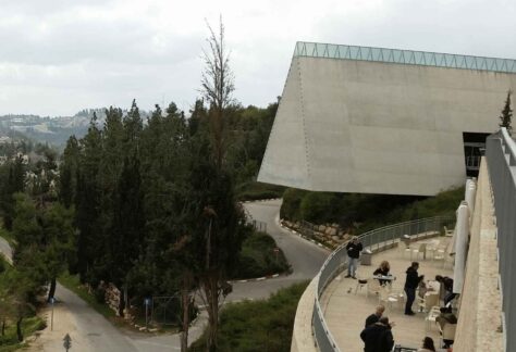 ein Gebäudekompplex auf einer Anhöhe. menschen befinden sich auf einer Terrasse mit Blick auf Tannen