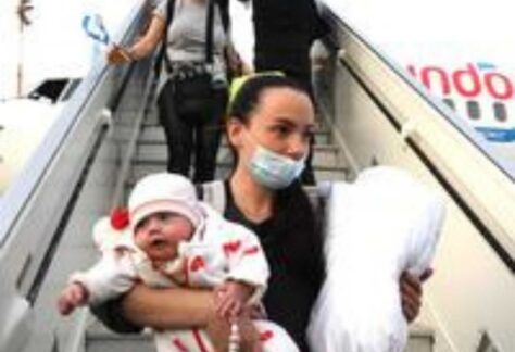 Menschen mit Mundschutzmaske steigen aus einem Flugzeud aus. Ganz vorne an eine Mutter mit ihrem Baby