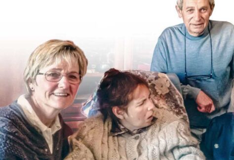 Eine Frau mit einer Behinderten Frau sitzend, ein Mann steht hinter den beiden