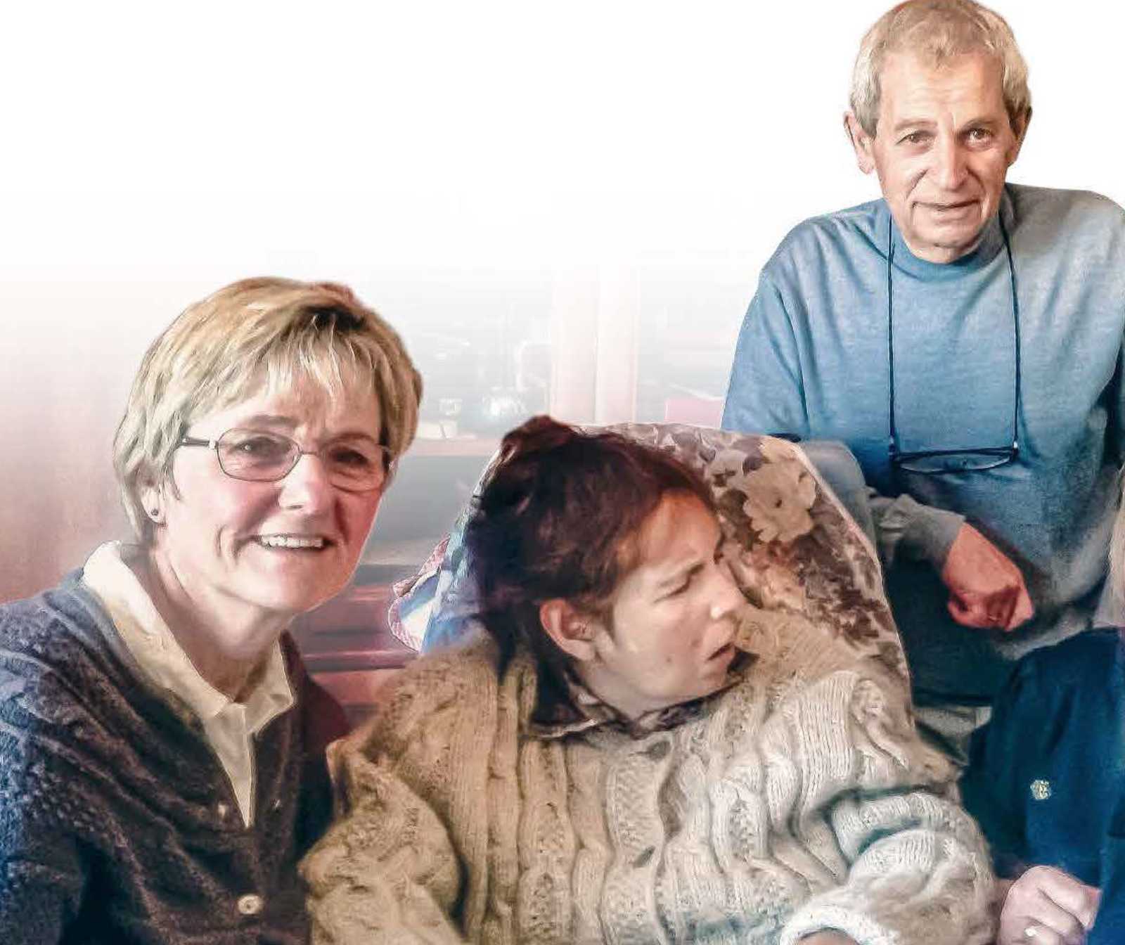 Eine Frau mit einer Behinderten Frau sitzend, ein Mann steht hinter den beiden