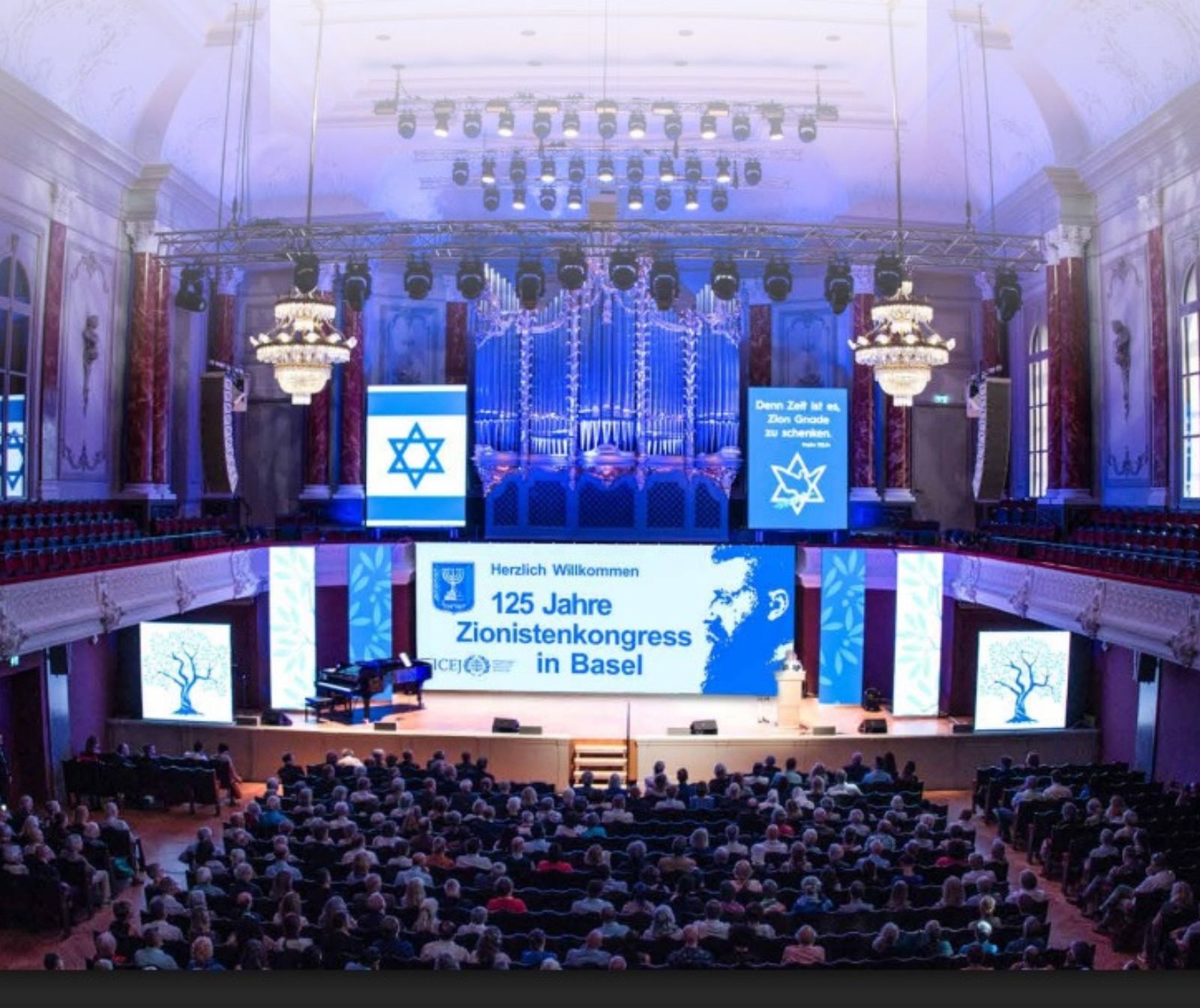 Blick vom Publikum aus auf eine Bühne, ein Schriftzug heißt Besucher auf dem 125. Zionistenkongress in Basel Willkommen. Ein Bildschirm zeigt die Flagge Israels, der Raum ist blau beleuchtet.