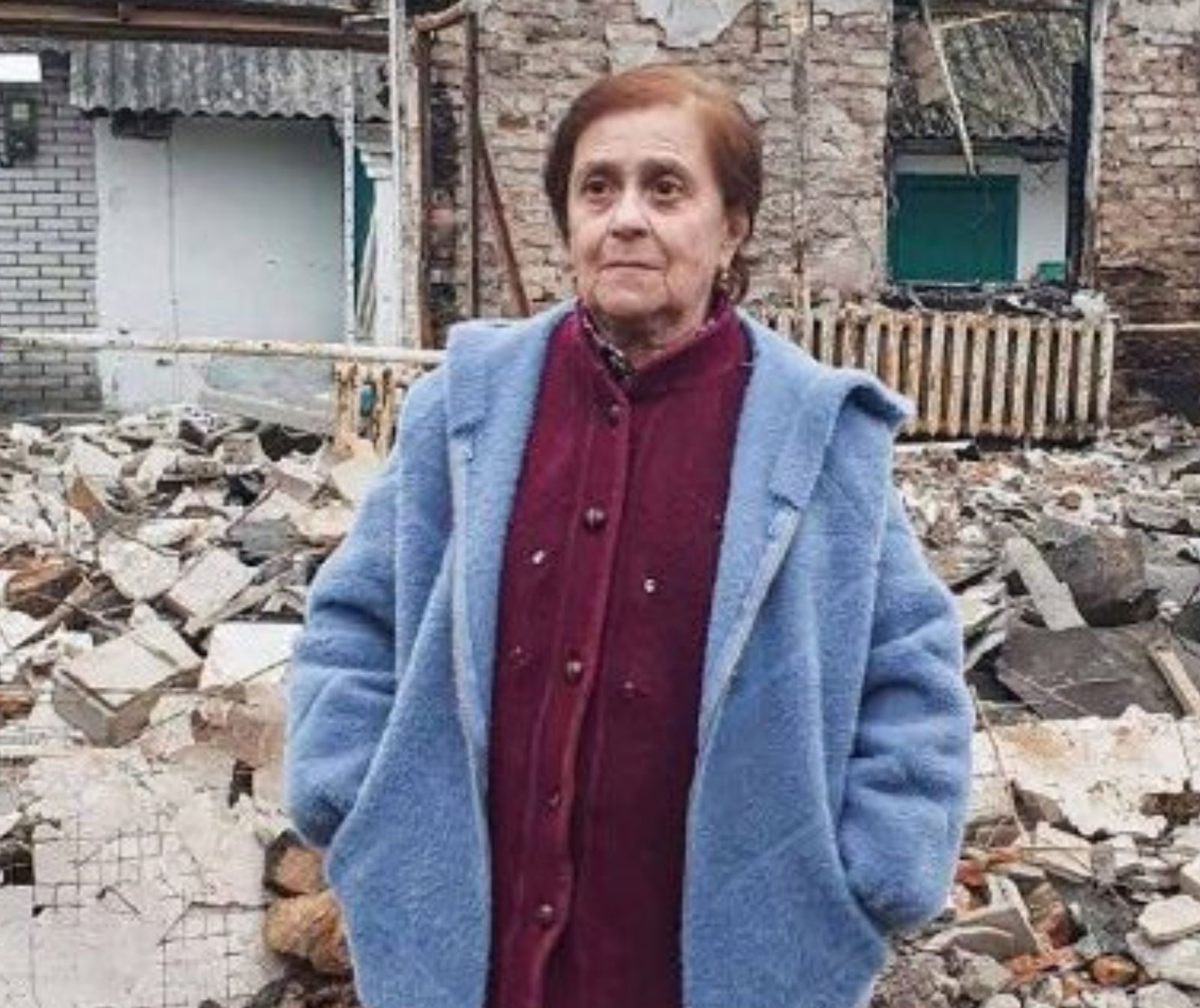 eine ältere Frau steht vor einem völlig zerstörtem Haus, sie trägt eine hellblaue Jacke