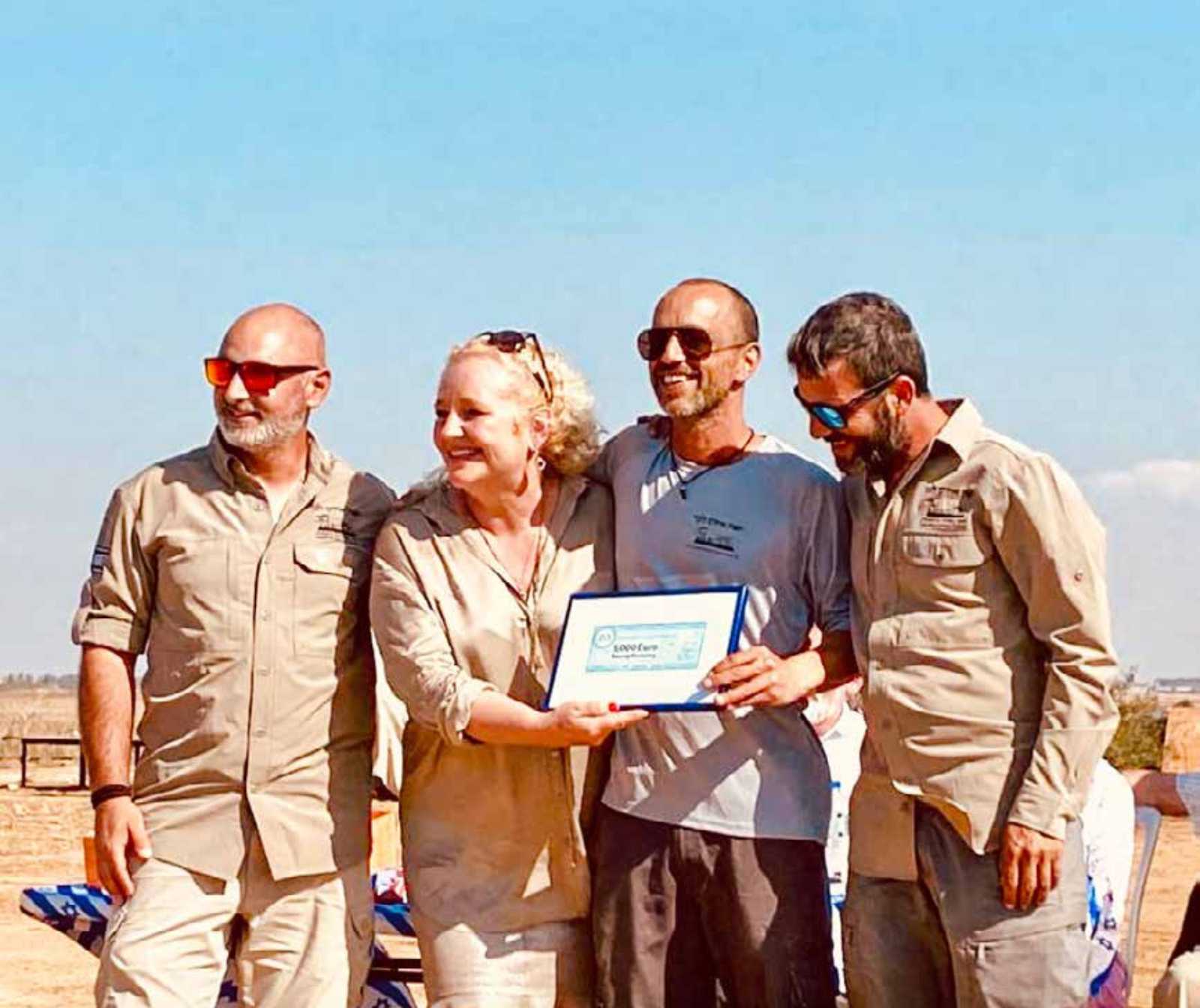 drei Männer und eine Frau in einer Reihe. Die zwei in der Mitte halten ein gerahmtes Dokument vor sich. Alle lächeln.