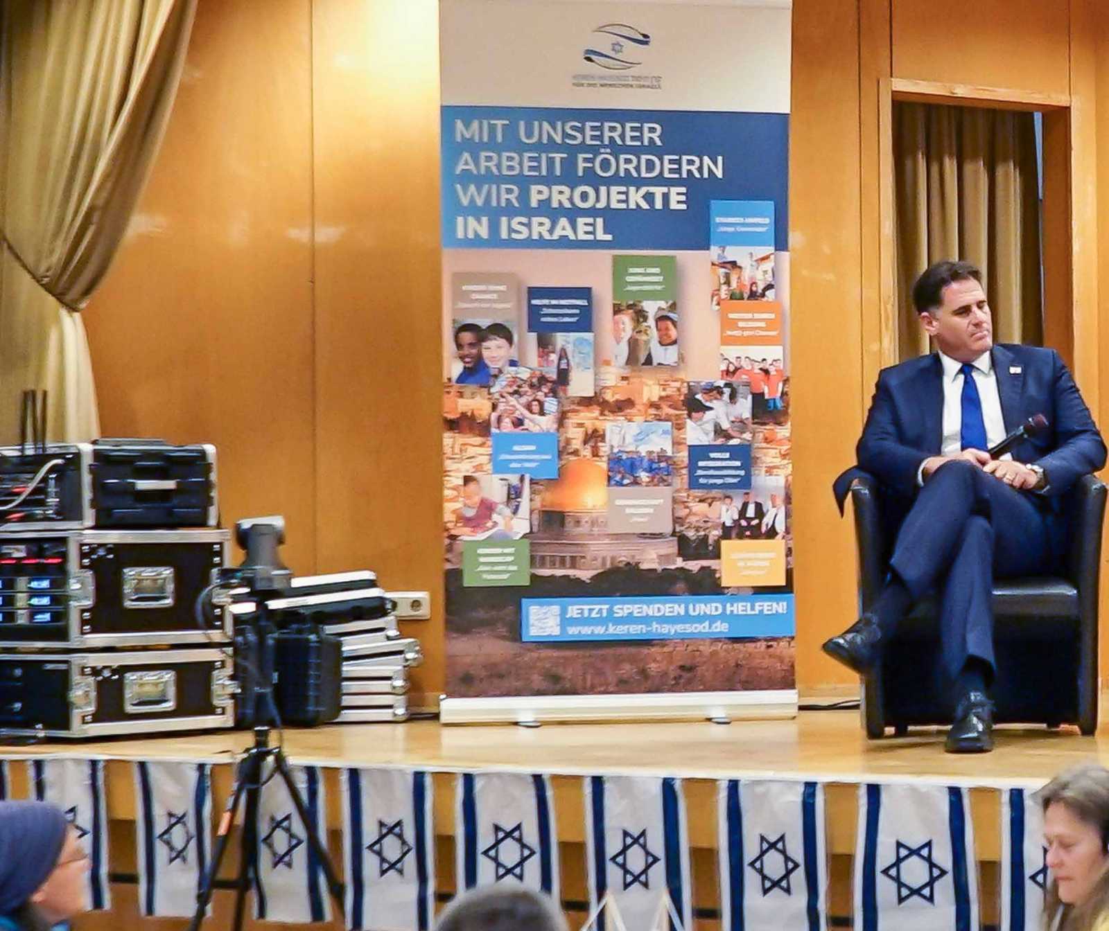Auf einer Bühne sitzt ein mann in Anzug. Neben ihm ein Banner, auf dem steht: Mit unserer Arbeit fördern wir Projekte in Israel. Vorne an der Bühne hängen Israelfahen nebeneinander hinunter.