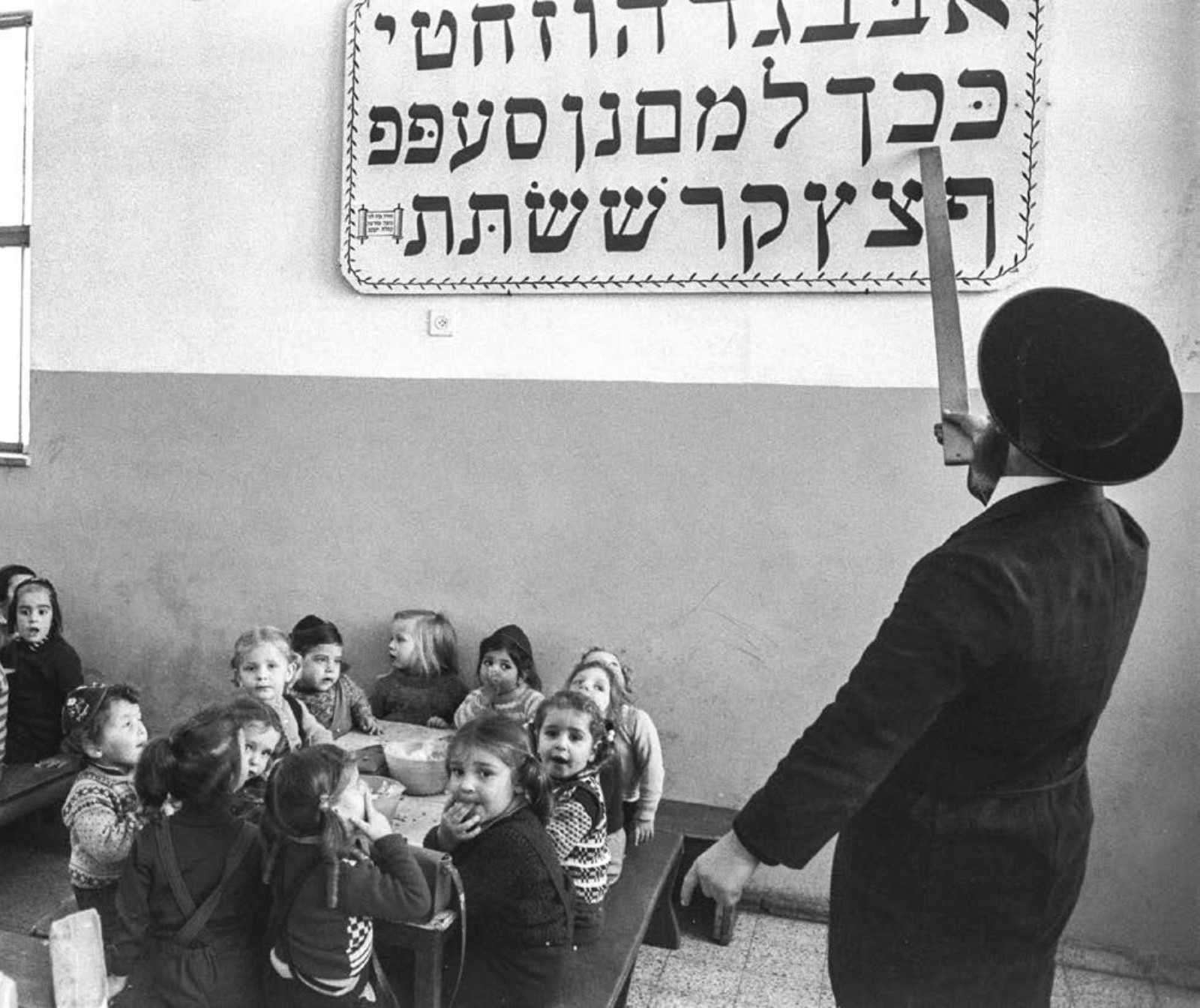 auf einem alten schwarz-weiß Foto ist ein Rabbi, der mit einem Stab auf eine an der Wand hängenden Tafel mit hebräischen Buchstaben zeigt. Vor ihm an einem Tisch sitzen kleine Kinder