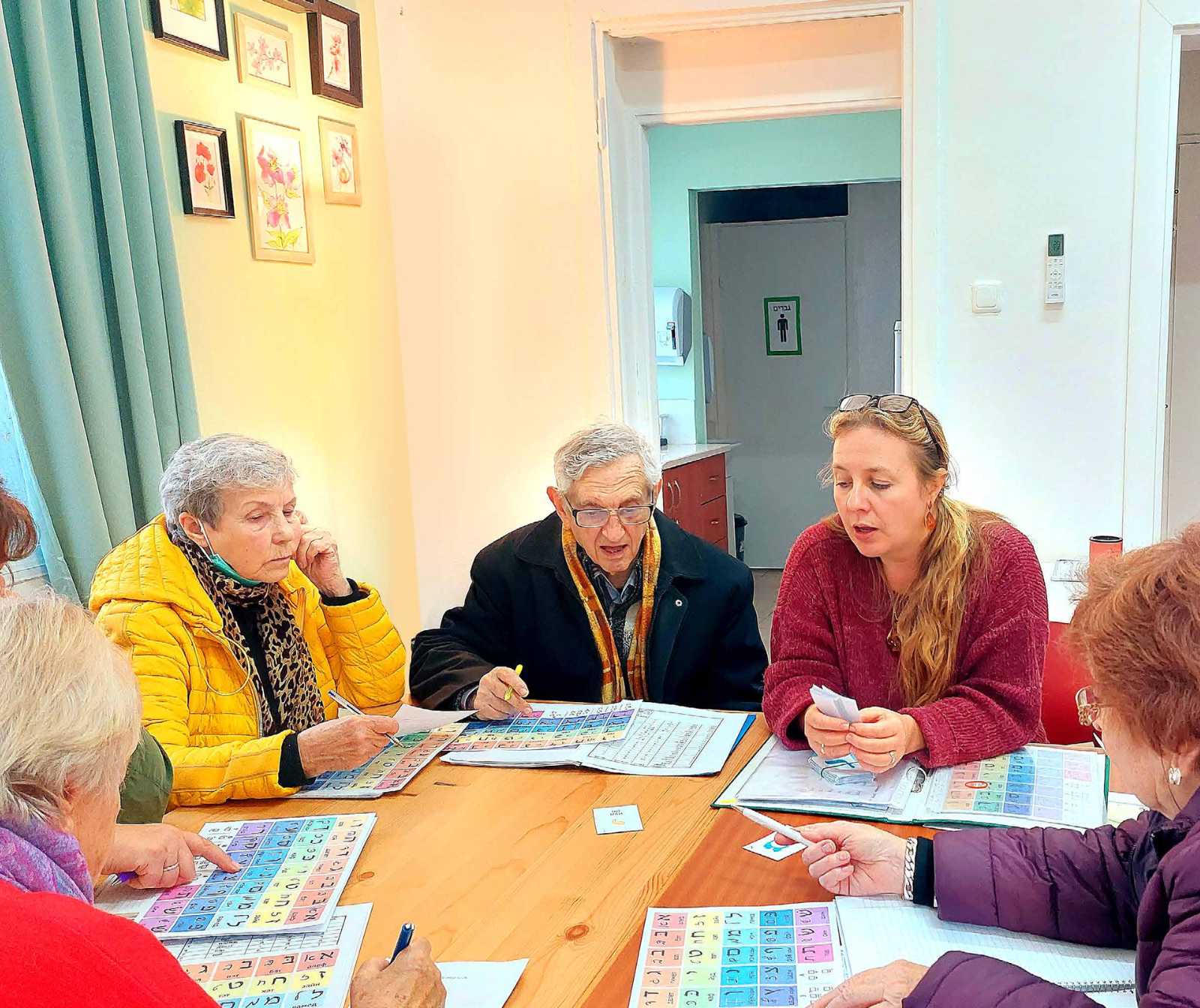 eine junge Frau sirtz mit mehreren älteren Herrschaften an einem Holztisch. Vor ihnen liegen Hefter mit bunten Schriftzeichen