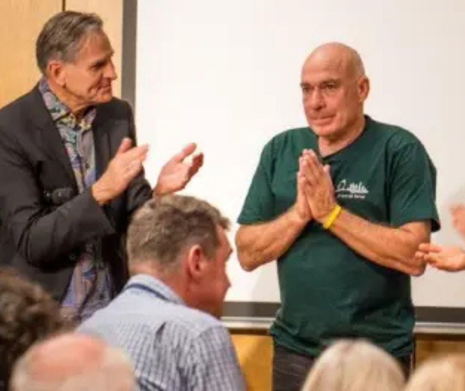 ein Mann mit grünem T-Shirt hat hält seine Hände, mit den Handflächen aneinander, vor seiner Brust, ein anderer mann klatscht Beifall