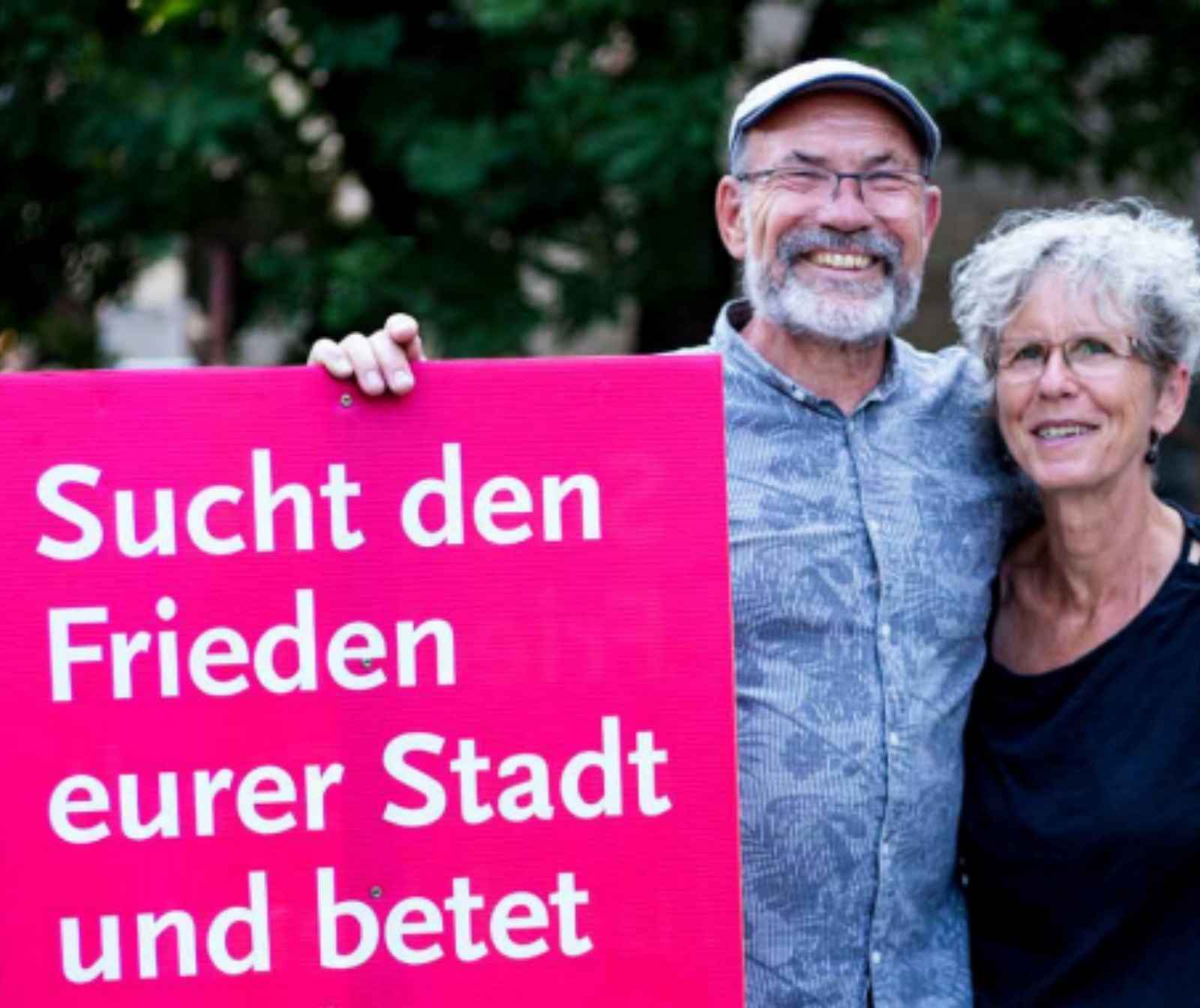 Ein Mann und eine Frau stehen nebeneinander. Der Arm des Mannes liegt um ihre Schulter. In der anderen Hand hält er ein Schild mit der Aufschrift "Sucht den Frieden in euerer Stadt und bete für sie zum HERRN.