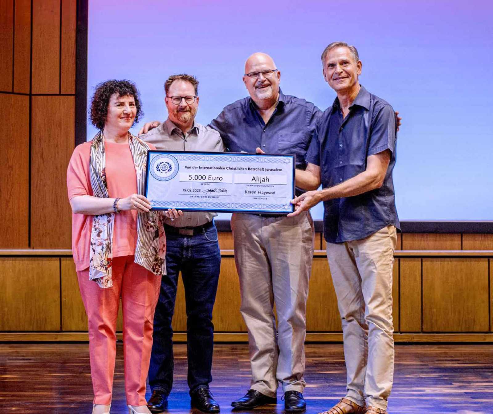 drei Männer und eine Frau stehen nebeneinander auf einer Bühne und halten einen großformatigen Scheck in der Hand. Sie lächeln