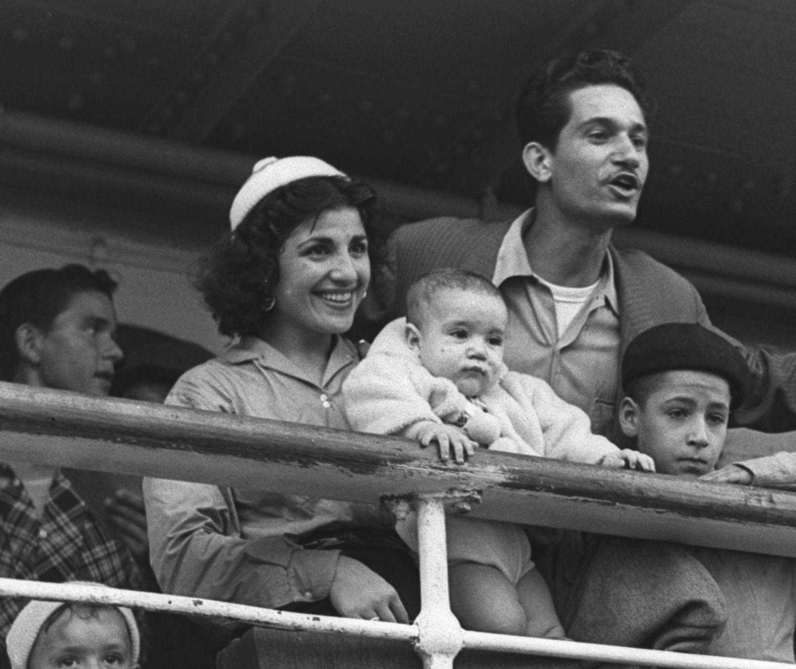 Mehrere Personen stehen an der Reling eines Schiffs. Links steht eine lächelnde Frau, die ein Kleinkind im Arm hält. Rechts hinter ihr lehnt sich ein Mann begeistert über die Reling, vor ihm steht ein Kind.