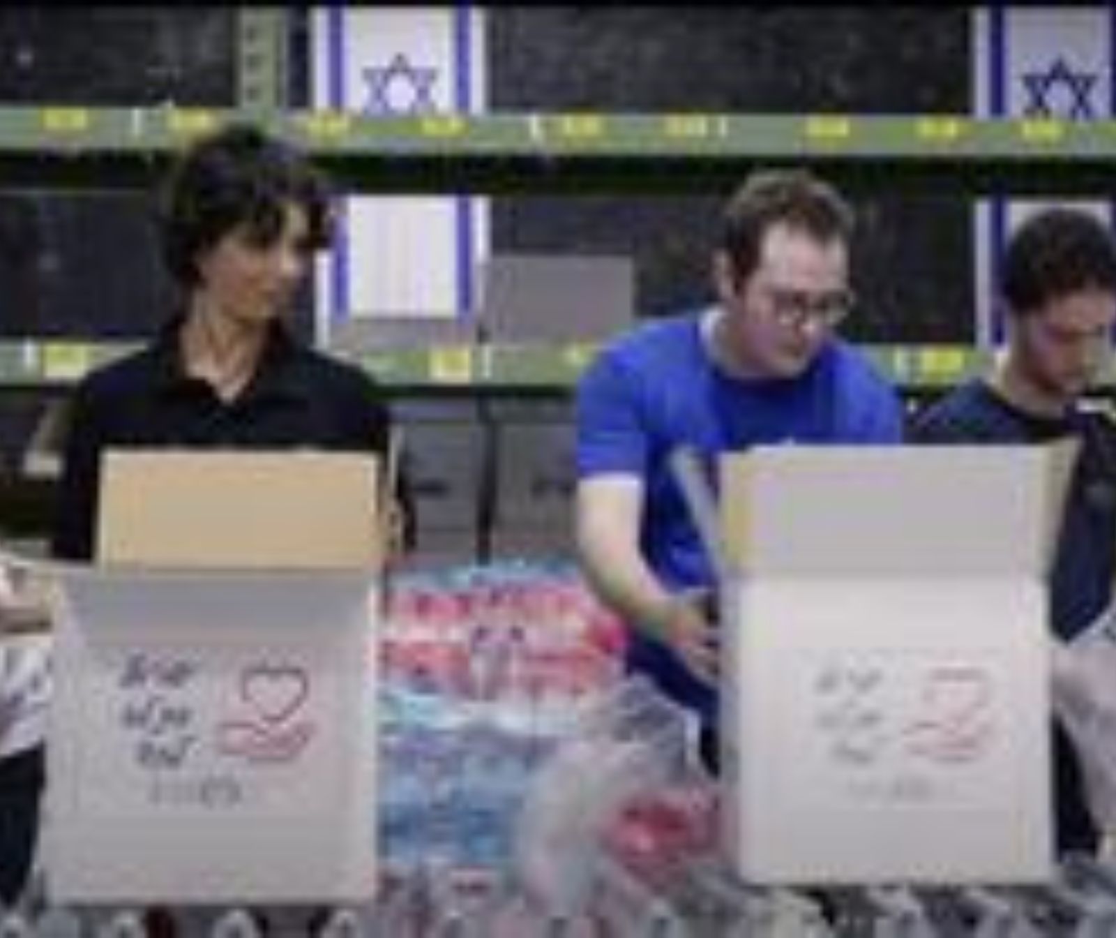 drei beschäftigte Menschen. Sie haben Kartons vor sich auf einem Tisch stehen. Im Hintergrund hängen zwei Israelfahnen