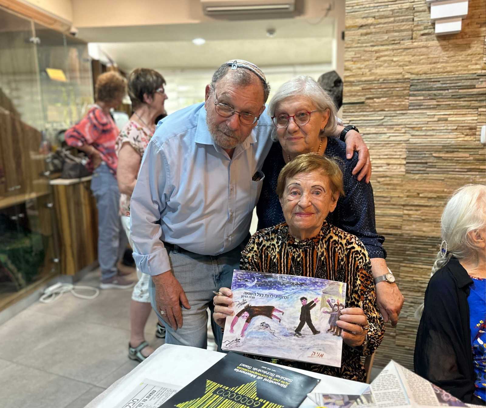 eine alte Frau zeigt ein Buch in die Kamera, hinter ihr stehen ein Mann mit Kipa und eine andere ältere Dame