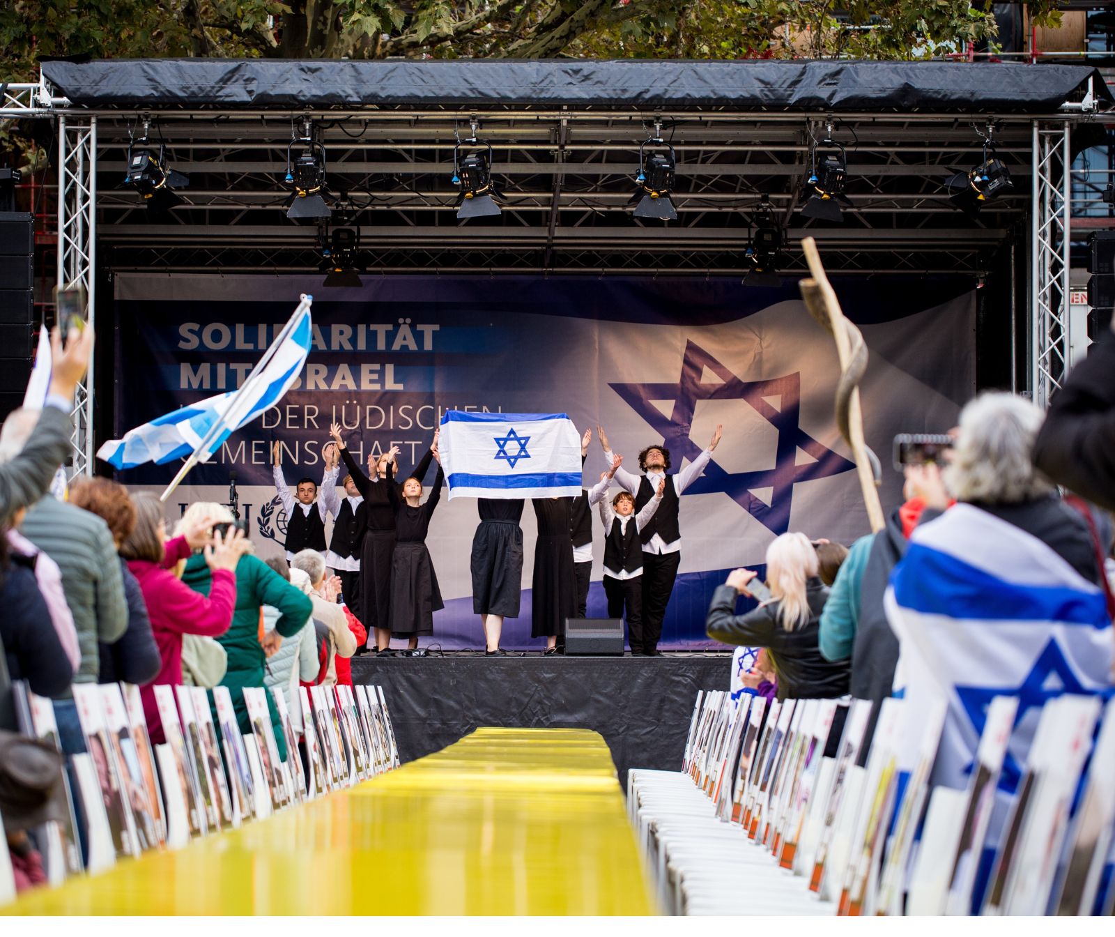 Auf einer Bühne steht eine Gruppe Kinder und Jugendliche in schwarzer Kleidung, sie halten eine Israelflagge. Vor der Bühne steht ein langer Tisch mit gelbem Tischtuch. Am Tisch stehen leere Stühle, an deren Lehnen die Bilder von Geiseln festgemacht sind.