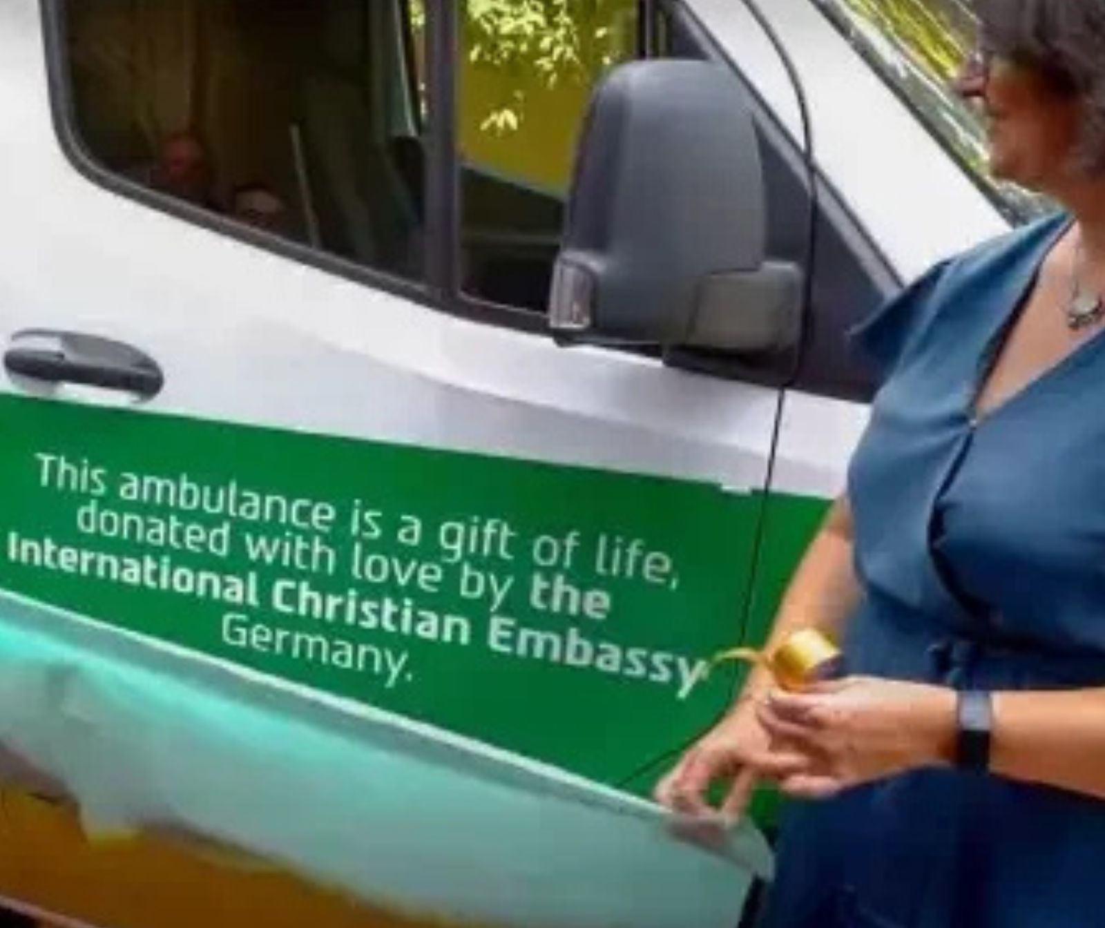eine Frau steht vor einem Transporter auf dem die Aufschriftsteht: This ambulance is a gift of life, donated with love by the International Christian Embassy Germany
