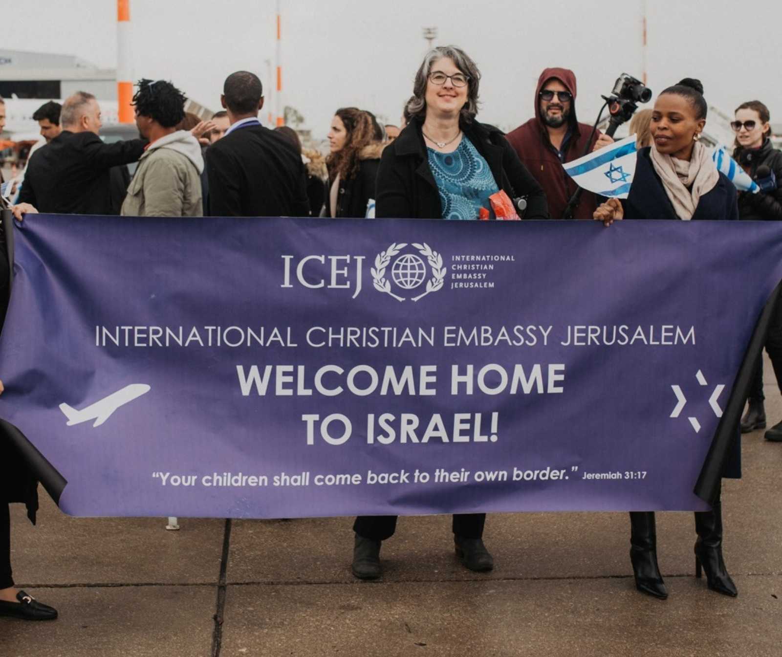 eine gruppe von menschen sind zu sehen, zwei frauen halten ein blaues banner mit ICEJ logo und der Aufschrift 'Welcome home to Israel' vor ihren Körpern