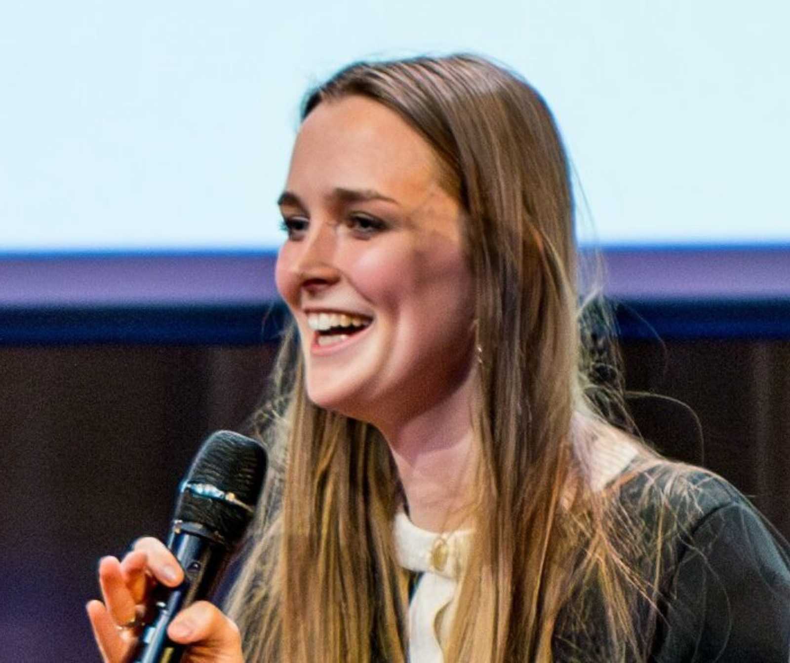 eine junge Frau mit langem blondem Haar hält ein Mikrofon in der rechten Hand. Sie lächelt.