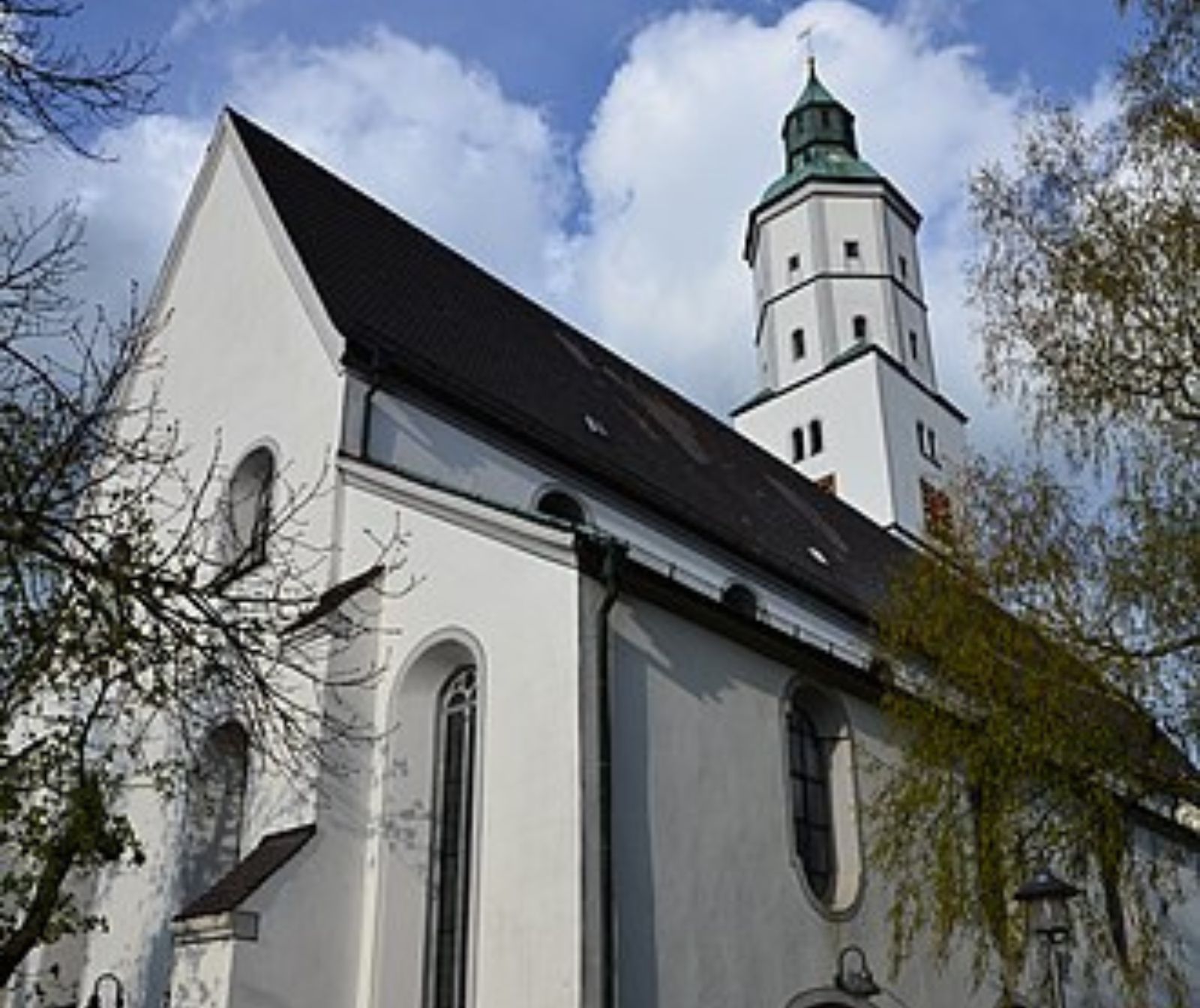 Umgeben von grünen Bäumen steht die weiße Kirche mit schwarzen Dachziegeln im Bildmittelpunkt. Vorne die Eingangstüre, im Hintergrund der Kirchturm mit grünem, spitzen Dach.
