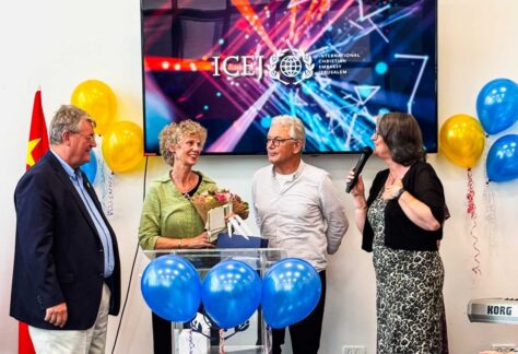 Gruppe von Menschen stehen vor einem Fernseher mit einem bunten Logo darauf, drumherum auch bunte Luftbalons