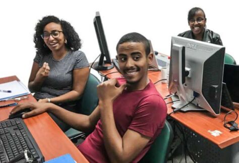 junge Menschen sitzen an ihren PCs an zwei Tischen und lächeln. Sie haben dunkle Hautfarbe