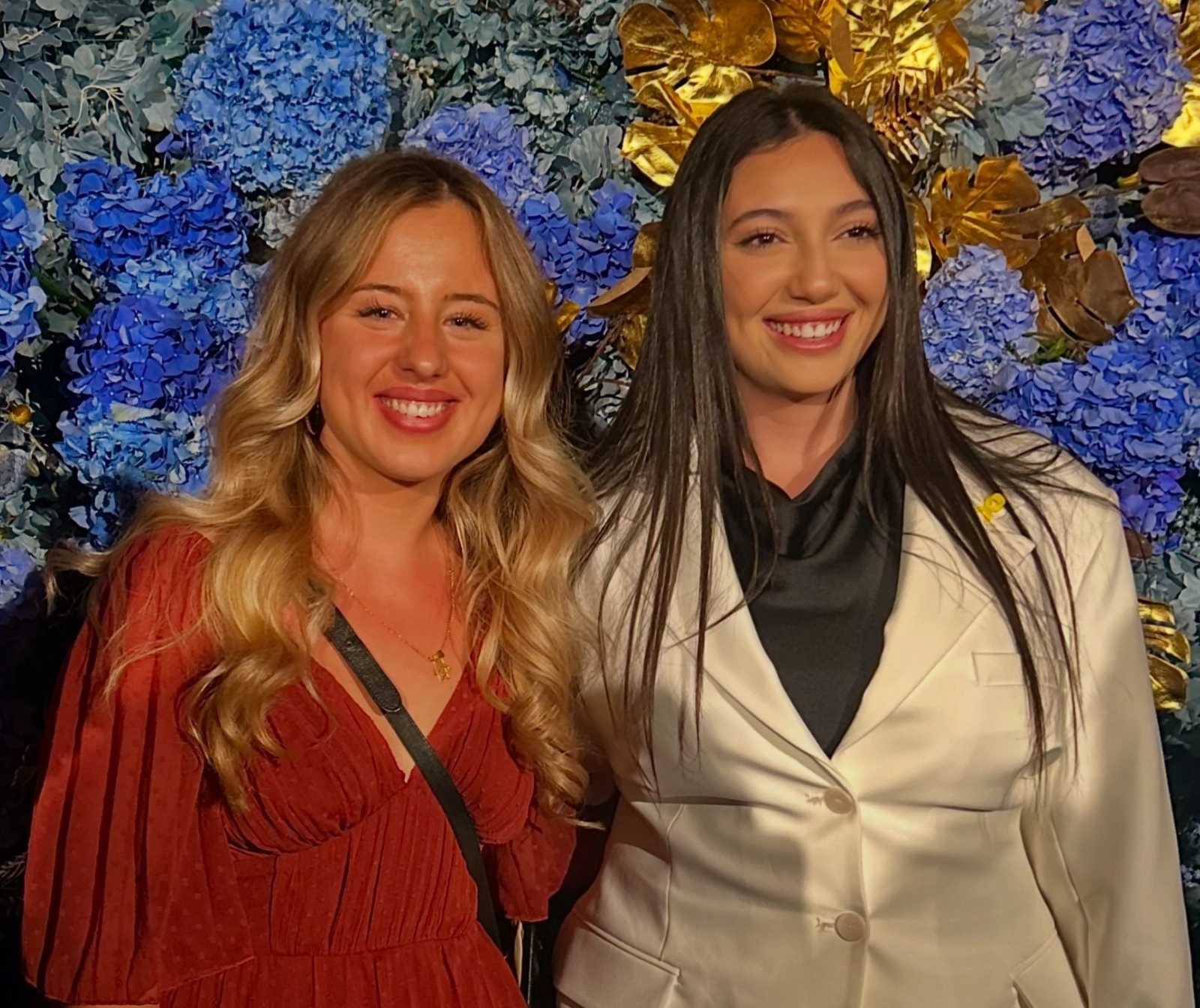 zwei jungen Frauen, eine blond, die andere dunkelhaarig, stehen eng nebeneinander und strahlen vor Freude