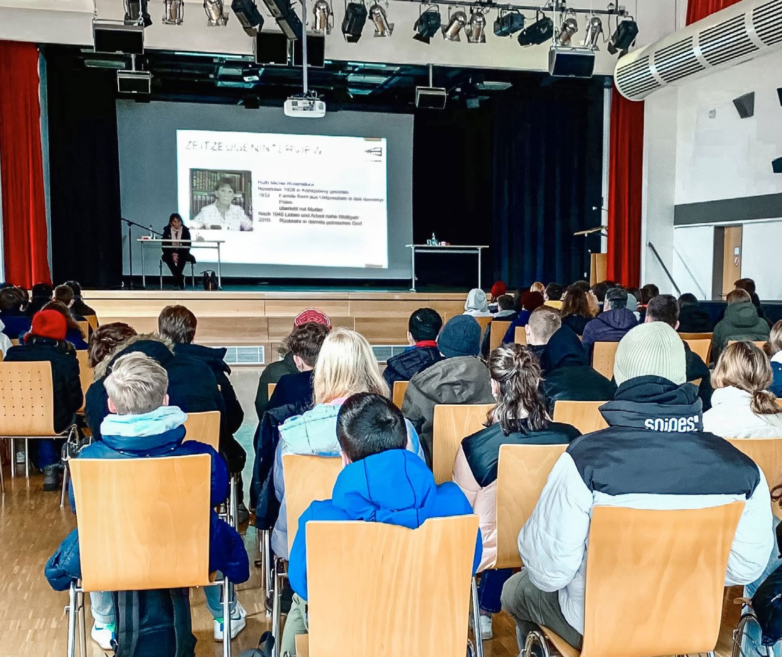 In einer Halle mit Bühne sitzen Menschen, vorrangig junge, in Jacken und sehen zur Bühne hoch. Dort sitzt eine Frau an einem Tisch, im Hintergruns ist etwas auf die Leinwand projeziert