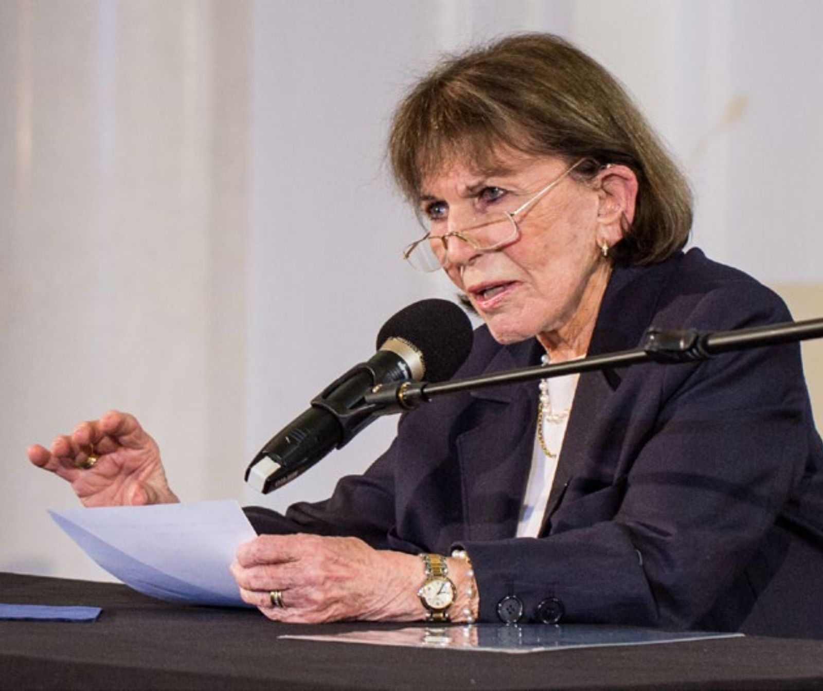 eine ältere Damen in schwarz gekleidet und einer Brille spricht an einem Tisch sitzend in ein Mikrofon. Sie hält dabei ein Blatt Papier in der Hand