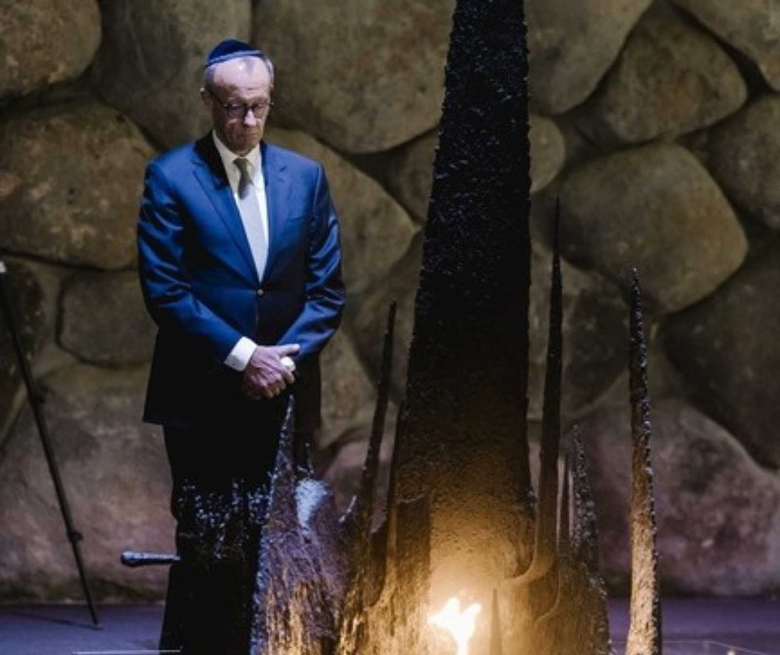 ein Mann in blauem Anzug, Brille und einer Kippa steht andächtig vor einer Skultur mit Feuer. Er hat den Kopf leicht nach unten geneigt und die Hände vor dem Bauch verschränkt
