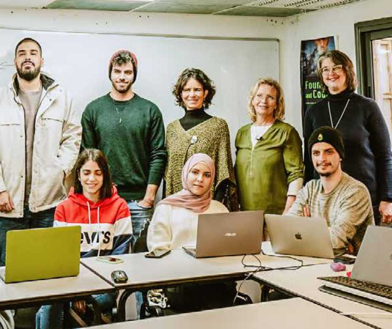 drei jüngere Menschen sitzen an Tischen, vor ihnen stehen PCs. Hinter ihnen stehen mehrere Personen