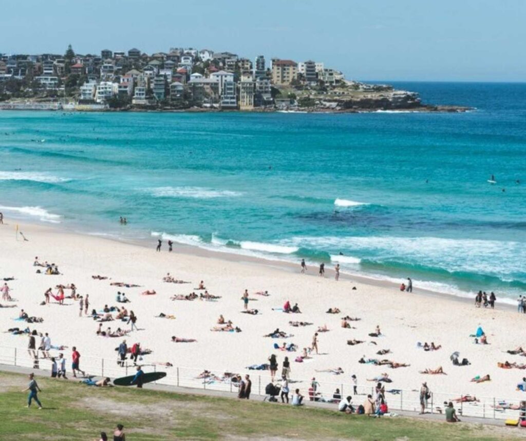 ein weißer Sandstrand mit Menschen belebt, türkisfarbenes Meer. Im Hintrgrund des Bildes eine Stadt
