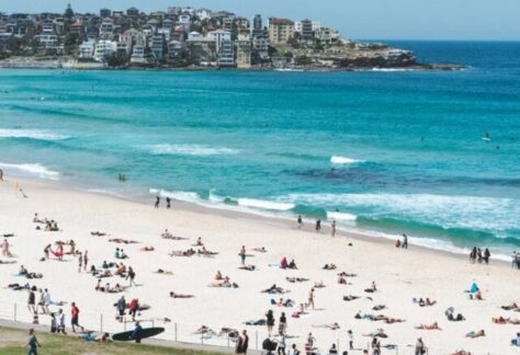 ein weißer Sandstrand mit Menschen belebt, türkisfarbenes Meer. Im Hintrgrund des Bildes eine Stadt