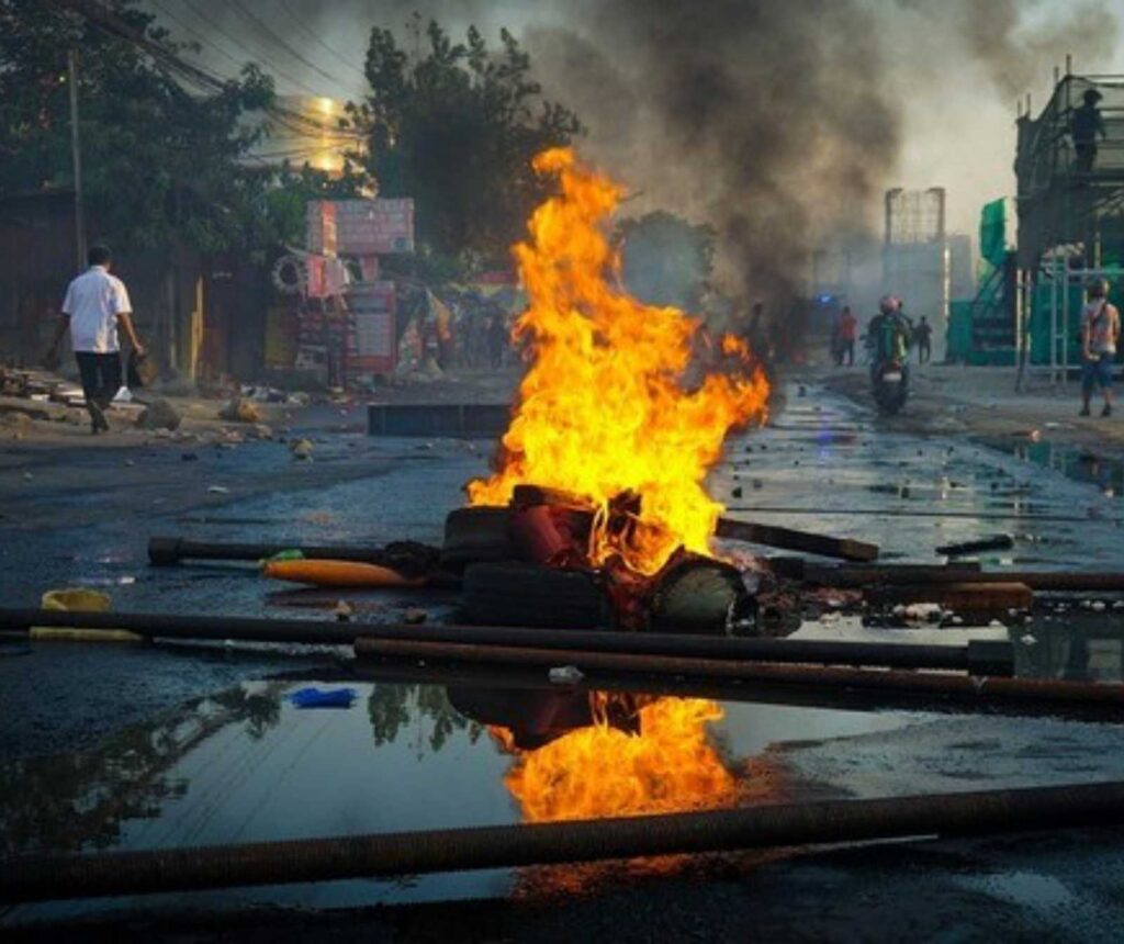 eine Straße auf der Dinge in Brand gesetzt sind. Im Hintergrund sind, Rauch steigt auf. Passanten zu sehen.