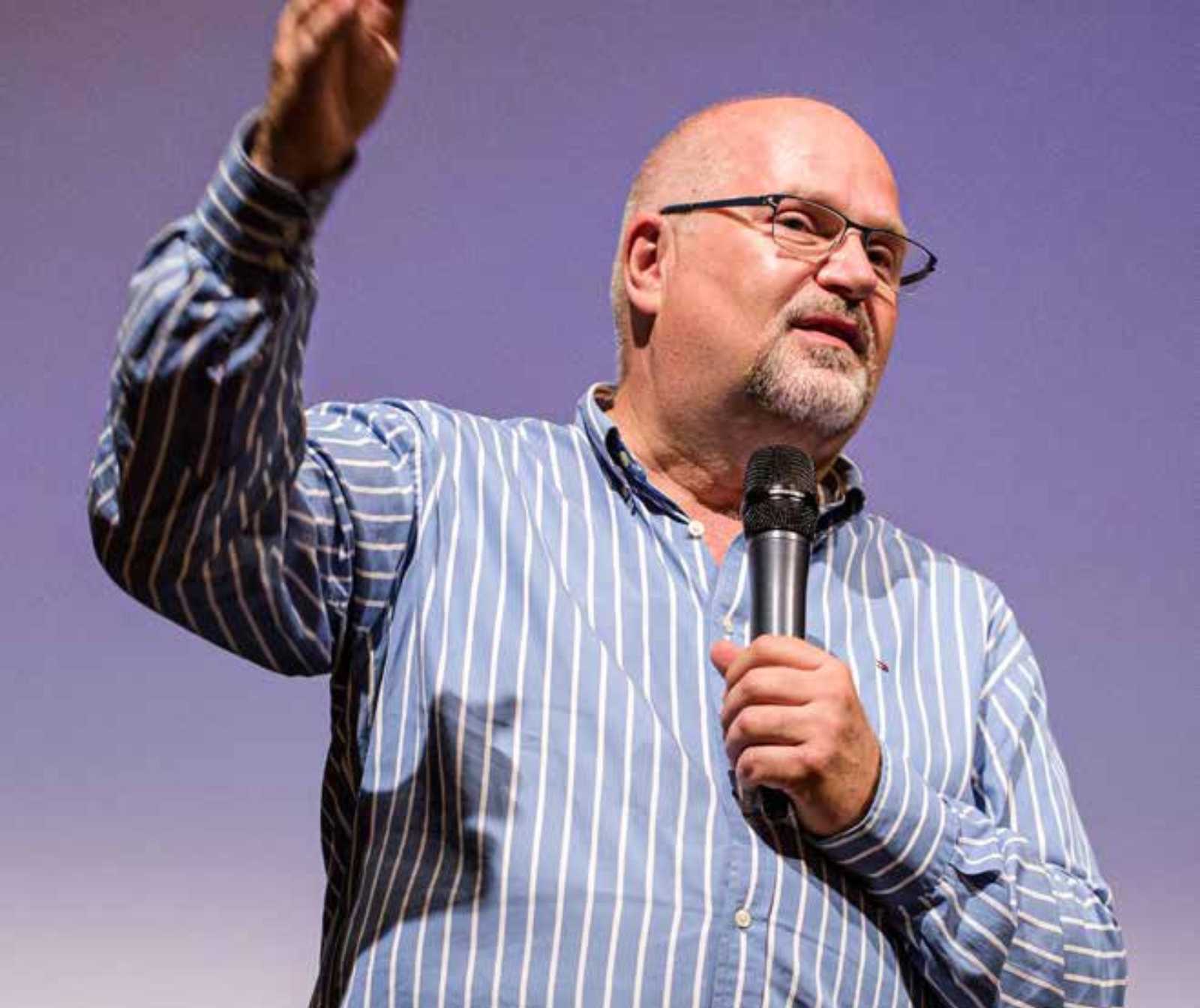 ein Mann mit gestreiftem Hemd und Brille, erhobener Hand und Mikrofon in der Linken Hand