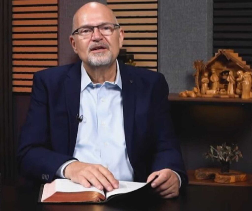 ein Mann mit blauem Anzug, weißem Hemd und Mikrofon am Jecket spricht während er ein Buch mit Goldrand auf dem Tisch vor sich auf dem Tisch liegen hat