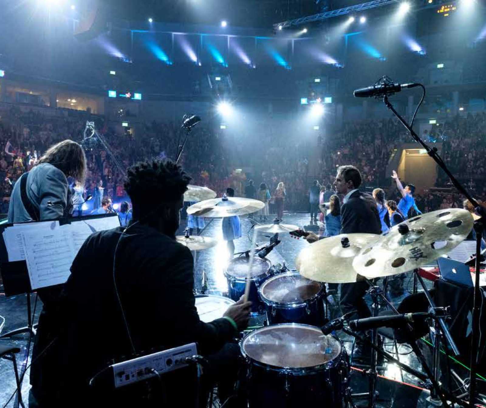eine Band auf einer großen Bühne in einem blau ausgeleuchtetem Saal. Der Blick geht hinter dem Schlagzeug in Richtung Publikum
