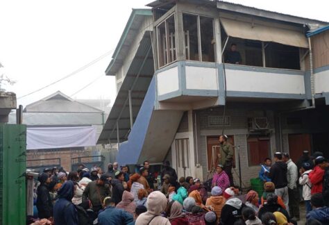 Vor einem Gebäude ist eine Menschenmenge versammelt. Die Menschen tragen warme Kleidung, sie haben eine dunklere Hautfarbe