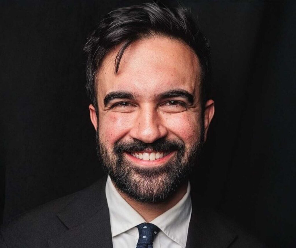 Headshot eines Mannes in schwarze, Anzug, weißem Hemd und Krawatte. Er lächelt