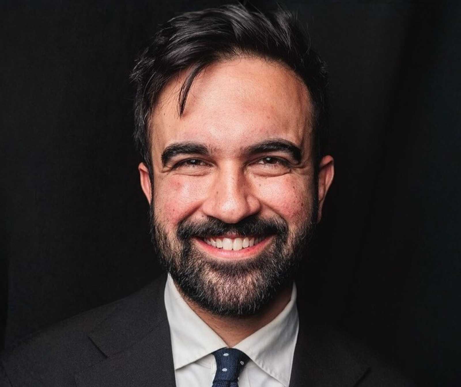 Headshot eines Mannes in schwarze, Anzug, weißem Hemd und Krawatte. Er lächelt