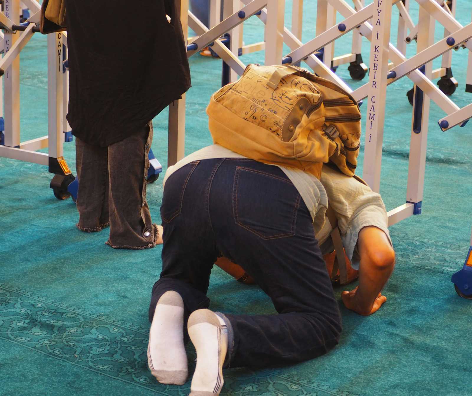 ein Mann mit Rucksack auf einem grünen teppich kniend, kopf auf dem Boden. Er trägt keine Schuhe