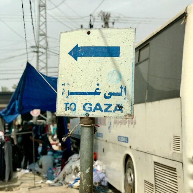 ein Schild mit blauer Aufschrift 'To Gaza' und ein Pfeil der nach links zeigt. Rechts im Bild ein Bus, im Hintergrund ein blaues Zelt