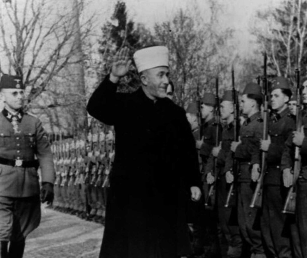 schwarz/weiß Fotografie eines Mannes mit traditioneller Kopfbedeckung. Er hebt seine rechte Hand zum Gruß, neben ihm Soldaten in Uniform, mit Gewehren
