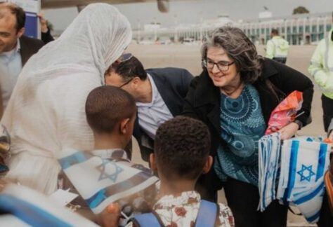 eine hellhäutige Frau mit Brille begrüßt eine Frau mit weißem Kopftuch und zwei dunkelhäutige Kinder. Ein junge trägt eine Israelfahne