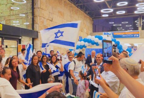 eine große Gruppe von Menschen unterschiedlichen Alters stehen strahlend in einer Halle beieinander. Viele tragen eine Israel Flagge. Es ist eine Girlande von blauen und weißen Luftballons aufgehängt