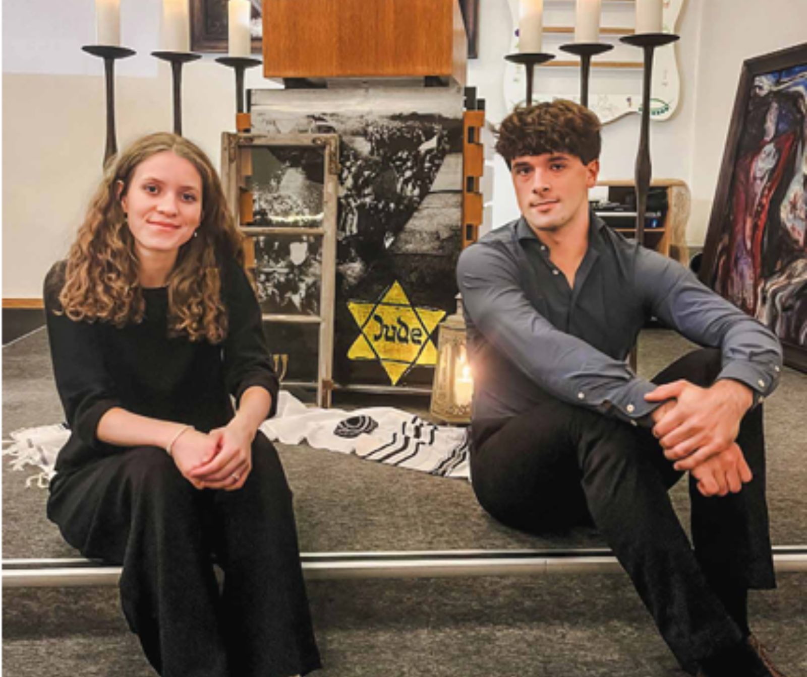 eine junge Frau und ein junger Mann sitzen nebeneinander auf einer Bühne und lächeln in die Kamera. Im Hintergrund sind Bilder Kerzenständer und ein Davidstern, auf dem 'Jude' steht