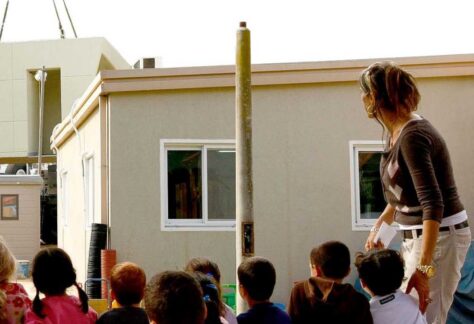 sitzende Kinder und eine stehende junge Frau schauen in eine Richtung, vor ihnen Containerartige Wohngebäude