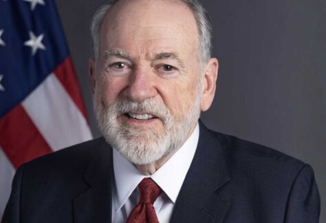 ein älterer Herr in blauem Anzug, weißen Hemd und roter Krawatte. Hinter ihm hängt eine USA Flagge