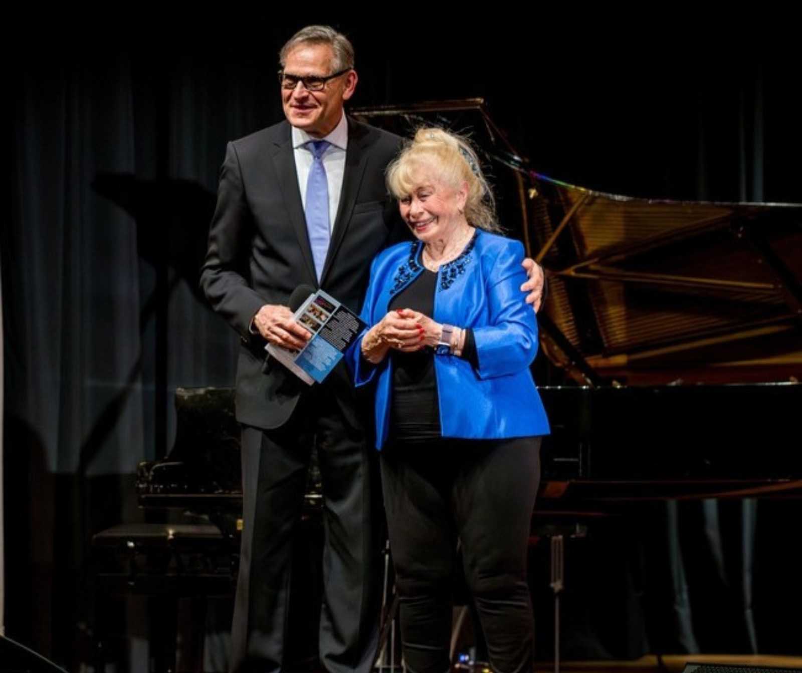 ein Mann in schwarzem Anzug und blauer Krawatte legt einer älteren Dame den Arm um die Schulter. Beide lächeln. Hinter ihnen steht ein Flügel